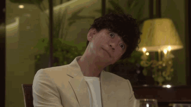 a man with curly hair is sitting at a table with a lamp in the background