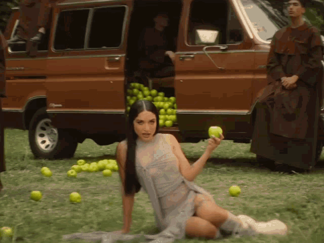 a woman in a sheer dress holds an apple in front of a brown van filled with apples