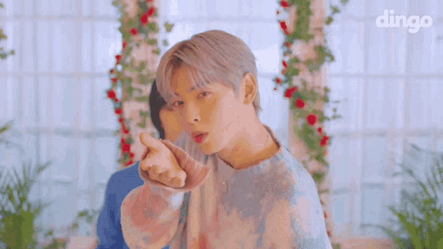a man in a tie dye sweater is pointing at the camera while standing in front of flowers .