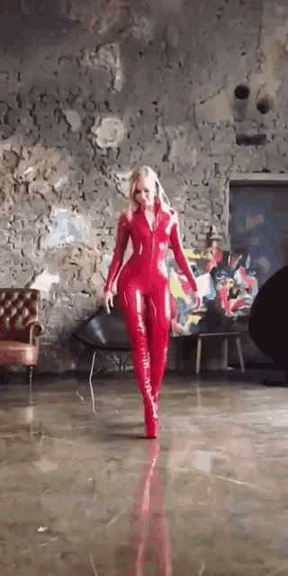 a woman in a red latex catsuit is standing in a room with a painting on the wall .
