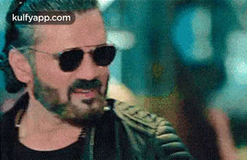 a man with a beard and sunglasses is wearing a leather jacket and a hat .