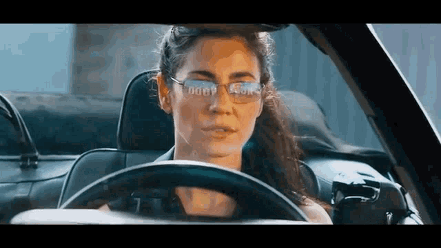 a woman wearing glasses is driving a car and looking at the camera .