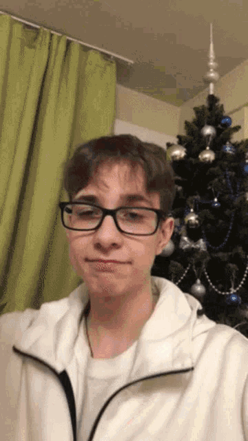 a young man wearing glasses and a white jacket stands in front of a christmas tree