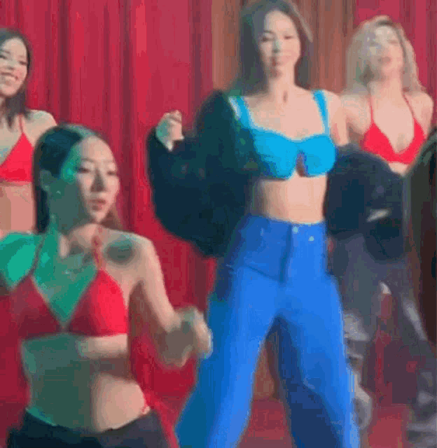 a group of women in bikini tops and blue pants are dancing on a stage in front of a red curtain .