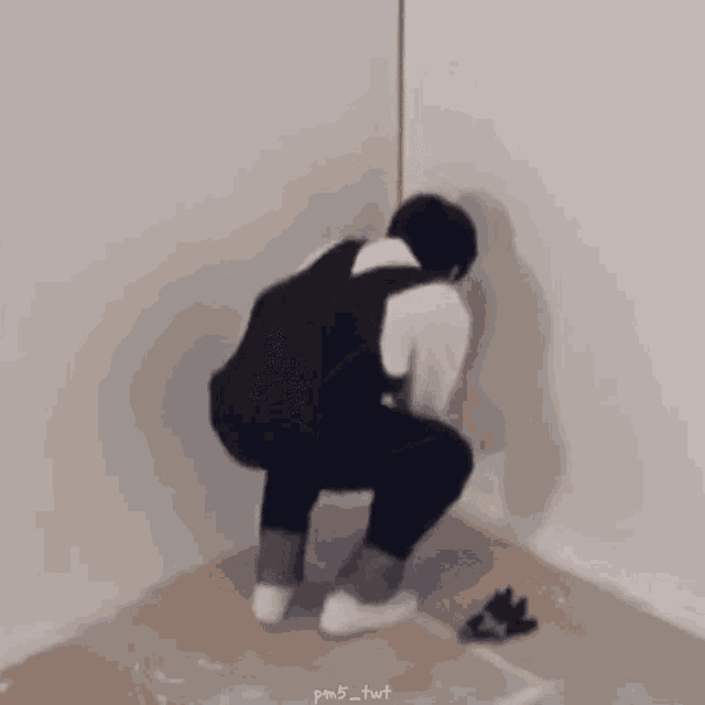 a man in overalls is squatting on the floor in a corner of a room .