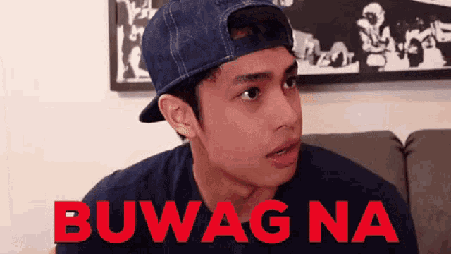 a young man wearing a baseball cap is sitting on a couch with the words buwag na written in red .