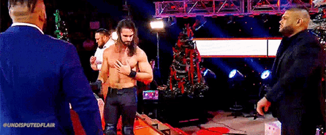 a man without a shirt is standing in front of a christmas tree on a stage with other men .