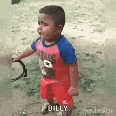 a little boy in a pink shirt and red shorts is standing in the dirt holding a rope .