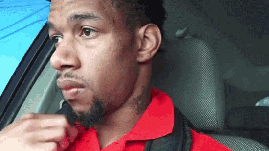 a man in a red shirt is sitting in the driver 's seat of a car looking out the window .