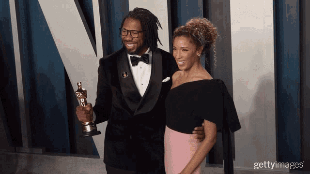 a man in a tuxedo is holding an oscar while standing next to a woman in a pink dress