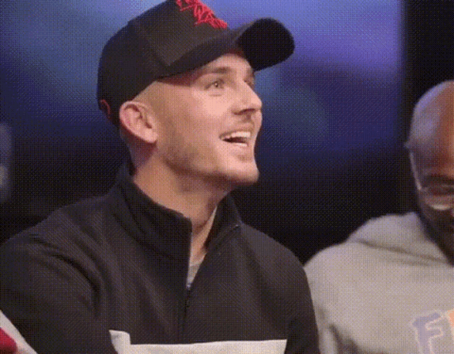 a man wearing a baseball cap and a black jacket is smiling while sitting next to another man .