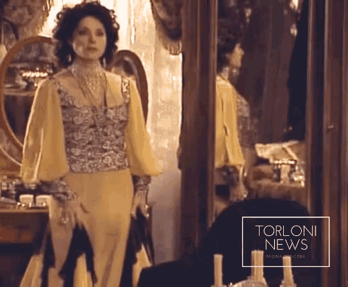 a woman in a yellow dress stands in front of a mirror with torloni news written on the corner