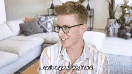 a man wearing glasses is sitting in a living room and says such a good boyfriend