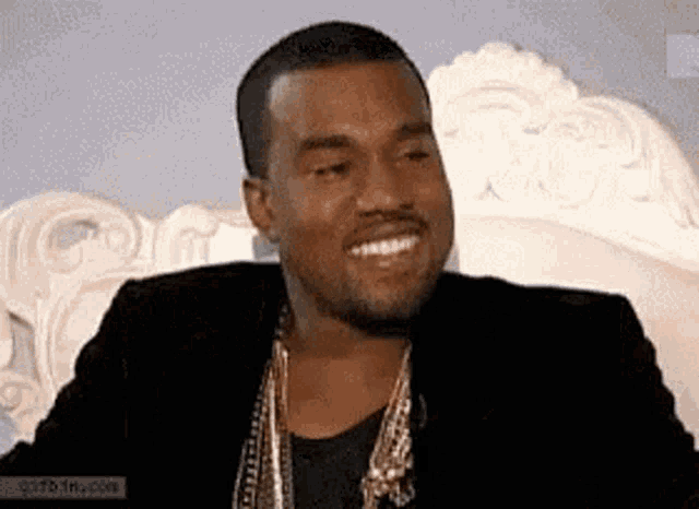 a man in a black jacket and gold necklace is smiling while sitting on a white couch .