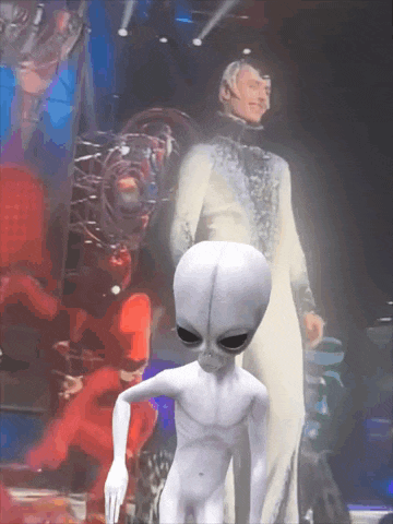 a white alien is standing on a stage with a man in a white suit behind him