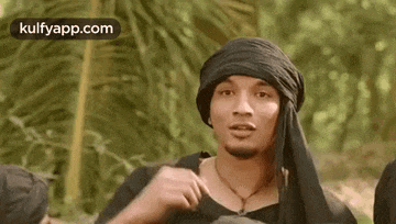 a man wearing a black turban and a black shirt is standing in a forest .