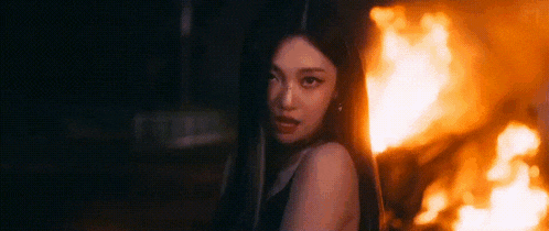 a woman is standing in front of a fire in the dark and looking at the camera .
