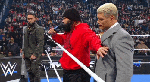 a man in a red hoodie is talking into a microphone while a man in a suit stands next to him