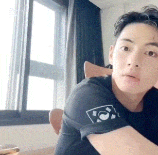 a young man is sitting in front of a window wearing a black shirt with a logo on the sleeve .