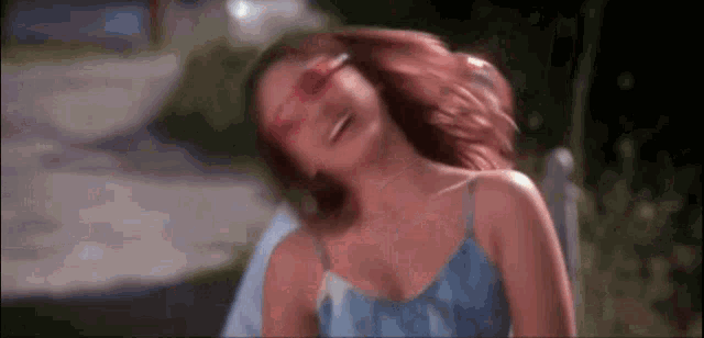 a woman wearing pink sunglasses is laughing with her hair blowing in the wind while sitting on a bench .