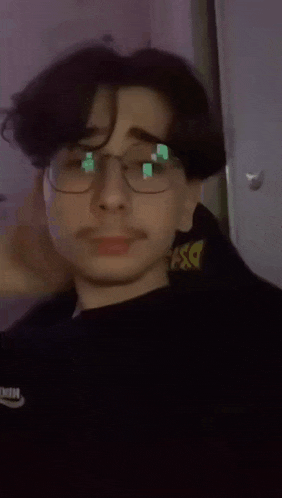 a man wearing glasses and a black sweater is covering his face with his hand .