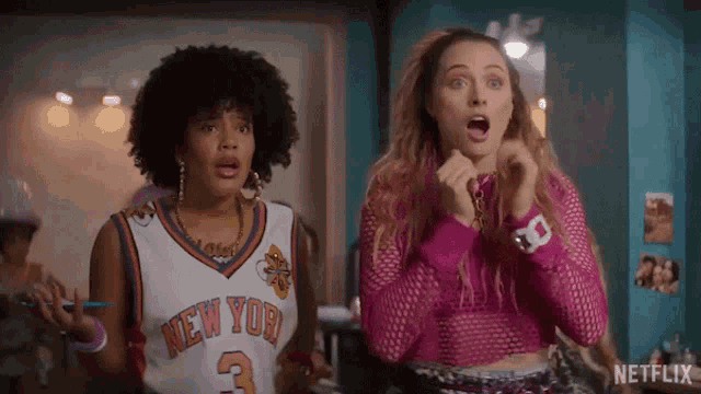 two women are standing next to each other in a room with their mouths open . one is wearing a new york jersey .