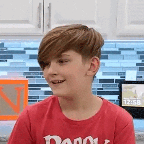 a young boy wearing a red t-shirt is smiling in front of a kitchen counter .