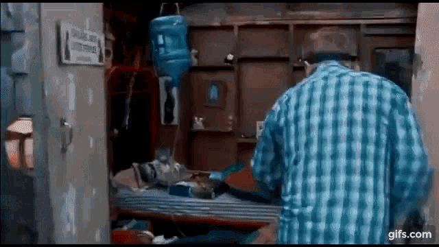 a man in a plaid shirt is standing in a room next to a bed with a water bottle hanging from the wall .