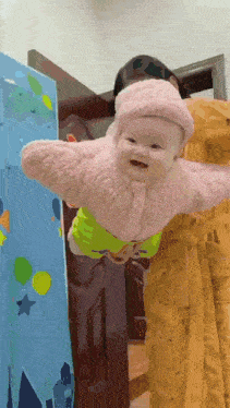 a baby in a pink jacket is being held by a woman in a yellow dress
