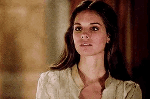 a woman with long brown hair is wearing a white shirt and earrings and looking at the camera .