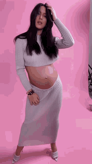 a pregnant woman is standing in front of a pink background wearing a crop top and a long skirt .