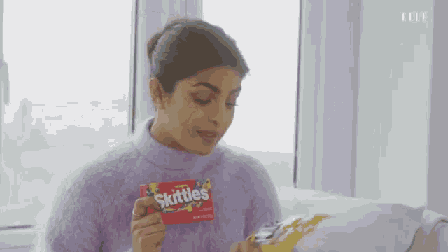 a woman in a purple sweater is holding a box of skittles and a bag of chips .