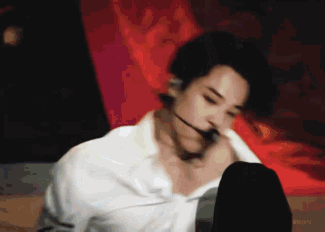a man in a white shirt is sitting on a bed with his legs crossed and a microphone in his mouth .