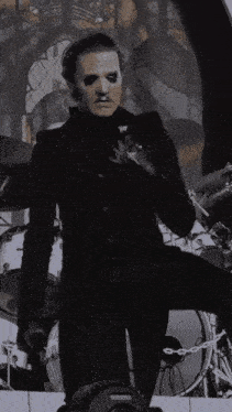 a man in a black suit is standing on a stage in front of a drum set and smoking a cigarette .
