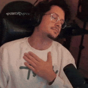 a man wearing glasses and headphones is sitting in front of a microphone with his hand on his chest .
