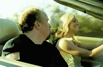 a man in a black shirt with an american flag on the back is driving a woman in a convertible .