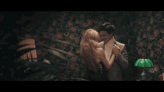 a woman with blonde hair is hugging a man in front of a floral wallpaper