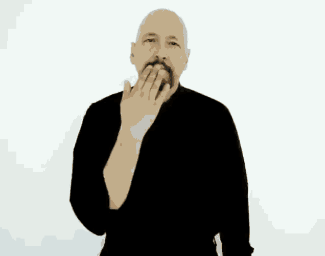 a bald man with a beard is wearing a black sweater and making a hand gesture .