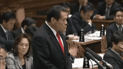 a man in a suit and red tie is giving a speech in front of a microphone in a parliament .