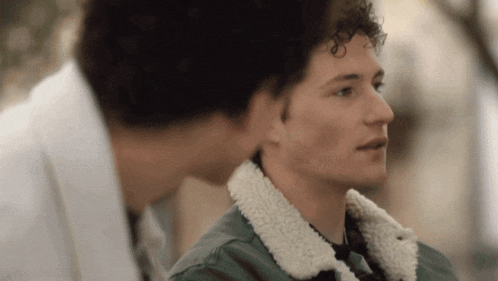 two young men are standing next to each other and looking at each other . one of the men is wearing a green jacket .