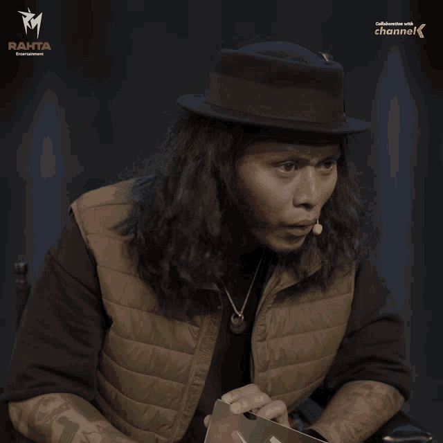 a man with long hair wearing a hat is sitting in front of a sign that says channel