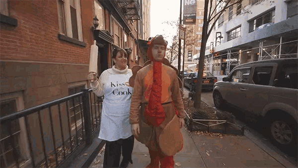a man in a turkey costume and a woman in a kiss cook apron walk down a sidewalk