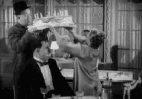 a black and white photo of a woman holding a cake over a man 's head in a restaurant .