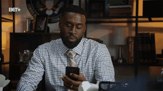a man in a plaid shirt and tie is looking at his phone with a bet logo in the corner