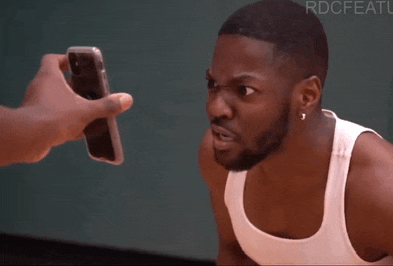 a man in a white tank top is looking at someone holding a cell phone in front of his face