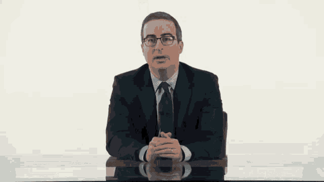 a man in a suit sits at a desk with his hands folded