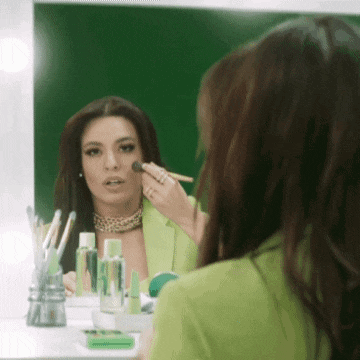 a woman in a green jacket applying makeup in front of a mirror