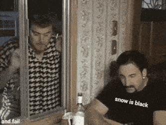 a man wearing a black shirt that says snow is black is sitting at a table with a bottle of alcohol .