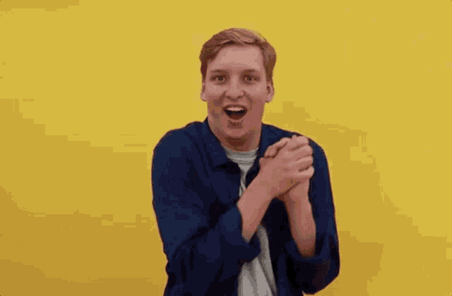 a young man in a blue shirt is making a funny face while standing in front of a yellow background .