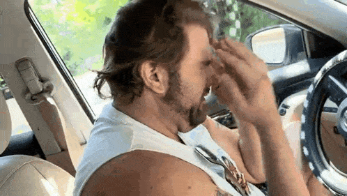 a man with a beard is sitting in the driver 's seat of a car covering his face with his hands .